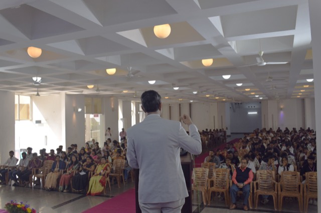 A speaker addresses a large audience of students from the floor