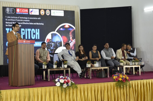 A speaker presents from the podium with the panel seated behind