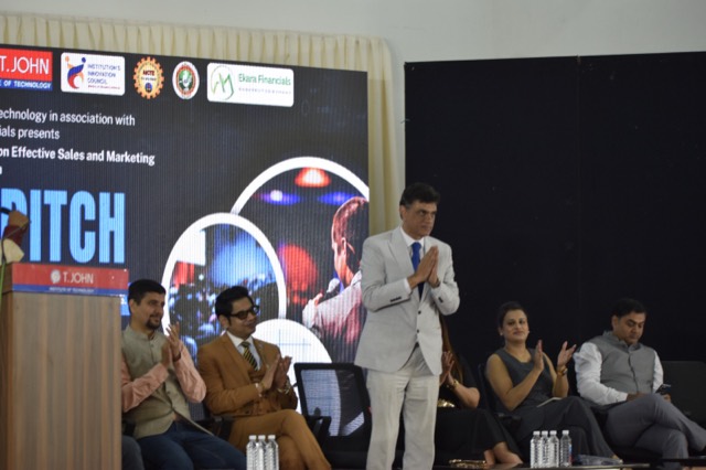 A guest greets the audience with folded hands beside the podium